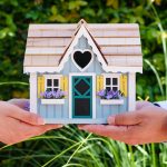 Two hands hold a colorful miniature house outdoors, symbolizing real estate and home ownership.